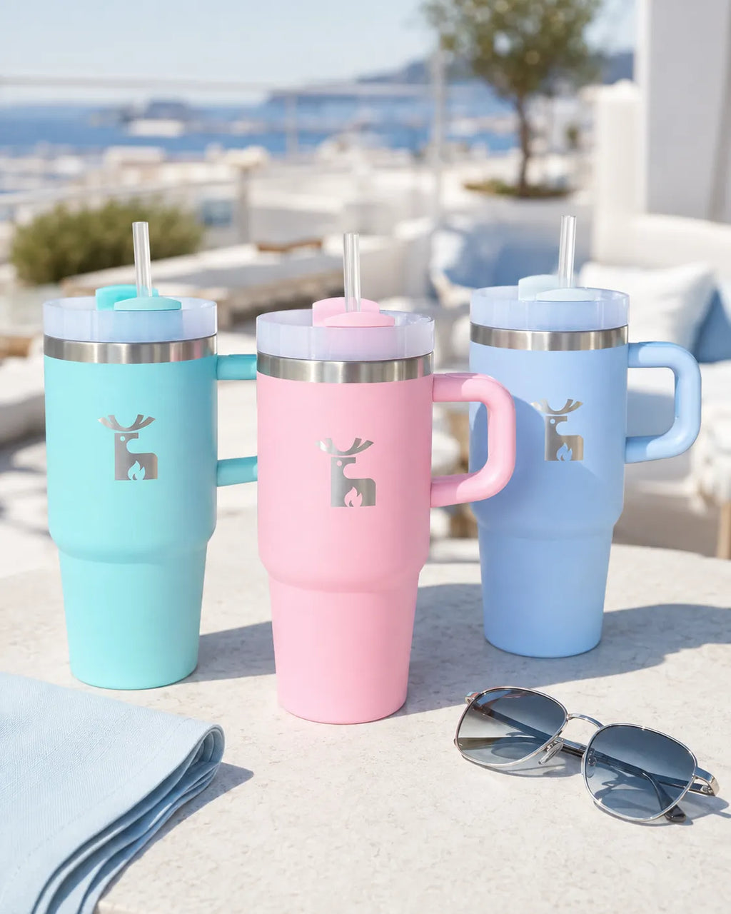 Three insulated travel tumblers with handles and straws in light blue, pastel pink, and mint green, arranged on a sunlit patio. Each tumbler has a silver deer logo on the front and a stainless steel rim.
