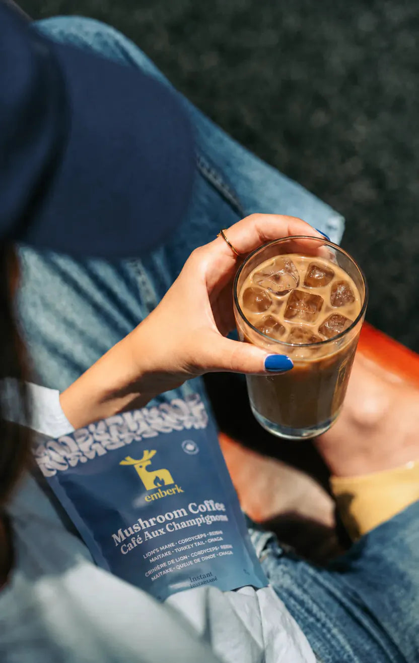 A person holding a glass of iced mushroom coffee with ice, next to a blue bag of Emberk Mushroom Coffee, while sitting on blue jeans.