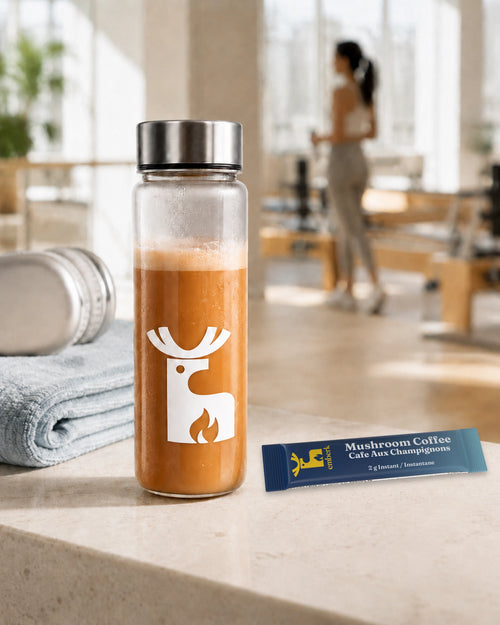 A clear glass bottle filled with mushroom coffee drink stands on a light stone surface in a bright indoor space. The bottle has a silver cap and a white deer logo with a flame detail.