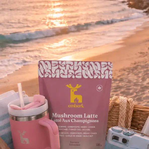 A pink package of Emberk Mushroom Latte is displayed on a beach picnic setup with a pink tumbler, a retro camera, and a wicker basket, set against a blurred sunset ocean background.