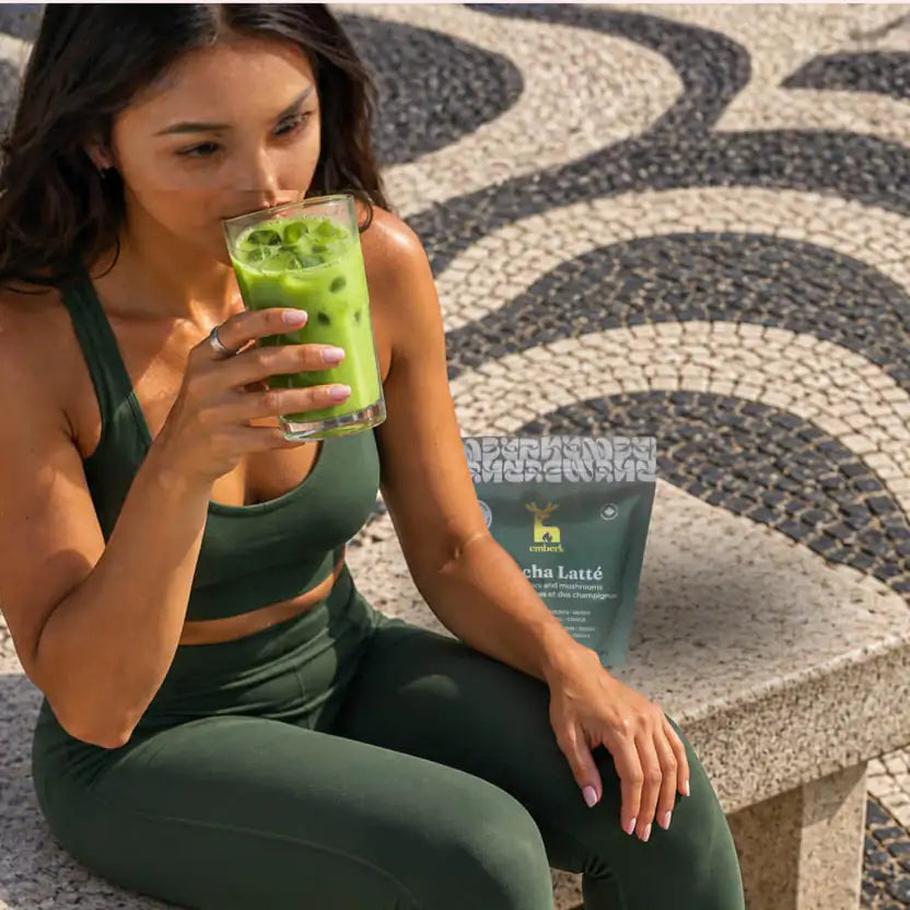 A woman in a dark green activewear set sits on a stone bench outdoors, holding a glass of matcha iced latte. A dark green pouch labeled 'Emberk Matcha Latte' with a deer logo rests beside her on the bench.